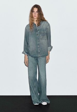 Jonge vrouw met lang golvend haar, gekleed in een oversized vervaagde denim shirt en losse denim broek, staande tegen een witte muur en een zwarte vloer.