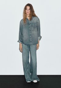 Young woman with long wavy hair wearing oversized faded denim shirt and loose denim pants standing against white wall and black floor.