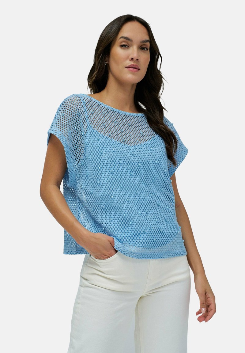 Top de malla suelta de color azul claro con cuello redondo y mangas cortas, que presenta un diseño texturizado de tejido abierto. Llevado con pantalones blancos.
