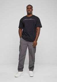 T-shirt en coton noir avec un logo script blanc, associé à un pantalon cargo gris avec des poches latérales et une coupe ample, complété par des baskets blanches.