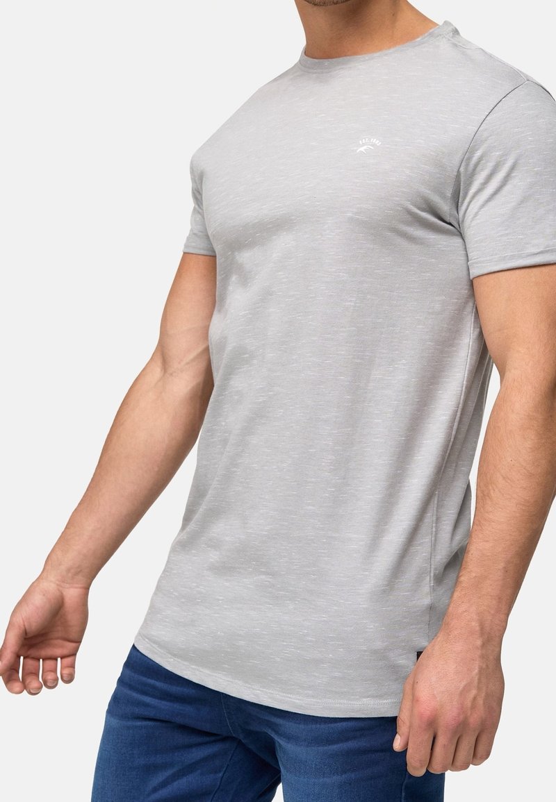 T-shirt gris à manches courtes en tissu léger avec une texture subtile, arborant un petit logo sur la poitrine. Porté avec un jean bleu.