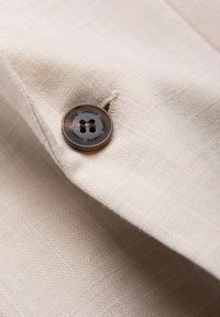 Bouton brun foncé avec le texte "OMBRE" cousu sur un tissu beige clair texturé, mettant en valeur le détail du tissu et la fixation du bouton.