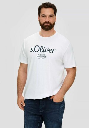 Bearded man wearing a white t-shirt with "s.Oliver Elevated Essentials Since 1969" text, standing with one hand in dark blue jeans pocket.