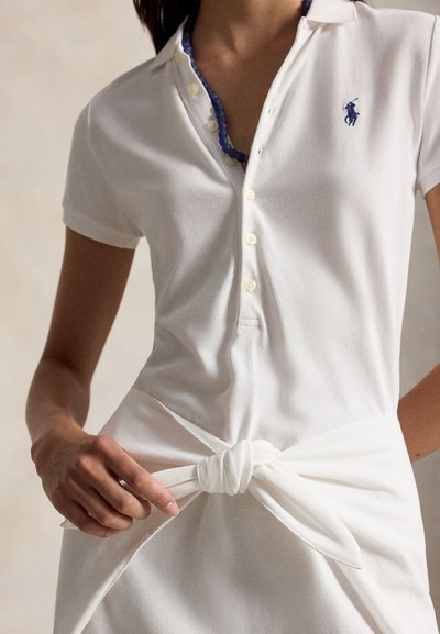 Nő fehér, rövid ujjú pöttyös dresszt visel, kék nyaklánccal és a deréknál megkötött textil övvel.