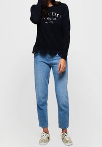 Mörkblå långärmad topp med spetsdetaljer och logotyp, matchad med ljusblå raka jeans och beige sneakers.