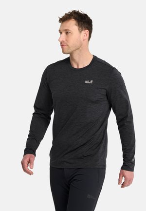 Mann mit kurzen lockigen Haaren, der ein dunkelgraues Langarmshirt und schwarze Hose trägt, schaut nach links vor einem schlichten Hintergrund.