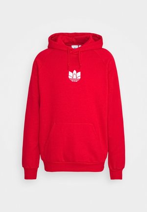 Mężczyzna w czerwonej bluzie z kapturem z białym logo, czarno-szarą kurtką w kratę, czarno-szarych spodniach z białymi logo oraz białych trampkach.