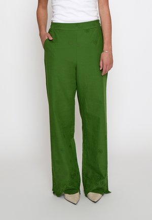 Pantalon vert à jambes larges avec des découpes florales œillet, porté avec un haut blanc et des chaussures beiges à bout pointu, les mains reposant sur les côtés.