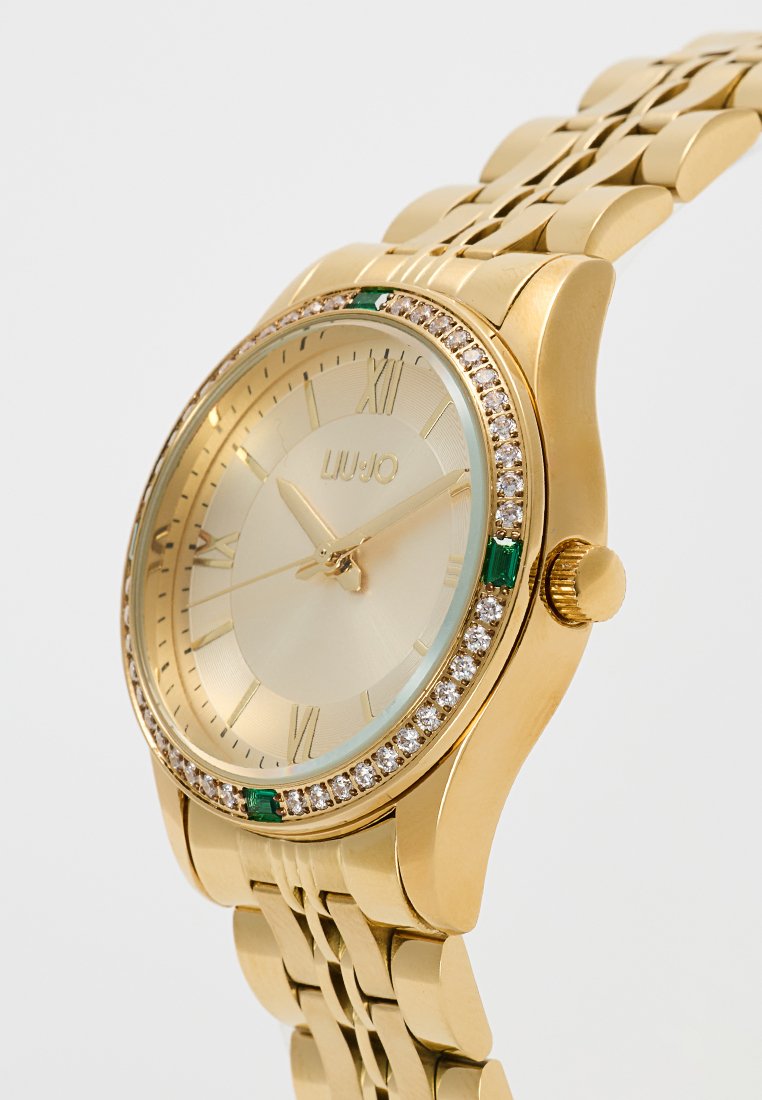 Gouden horloge met een ronde kast, een structuurdial en Romeinse cijfers. Heeft een met kristallen bezette bezel en groene accenten aan de zijkanten.