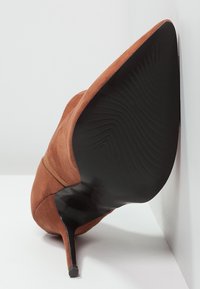 Tacchi alti in suede marrone con punta affilata e suola nera elegante. Presentano una superficie liscia e una curva sottile alla base del tacco.