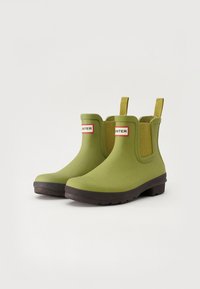 Botas de goma verdes hasta el tobillo con una suela negra, que cuentan con paneles laterales elásticos y una etiqueta de logo en rojo y blanco en la parte delantera. Superficie texturizada.
