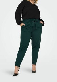 Woman wearing dark green high-waist pants with a waist tie, black blouse with lace neckline, and black high-heeled shoes, standing with hands in pockets.