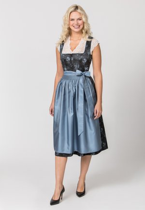 Lächelnde Frau, die ein schwarzes Dirndl mit Blumenmuster, eine weiße Spitzenbluse und eine blaue Schürze trägt, steht vor einem einfarbigen hellen Hintergrund.