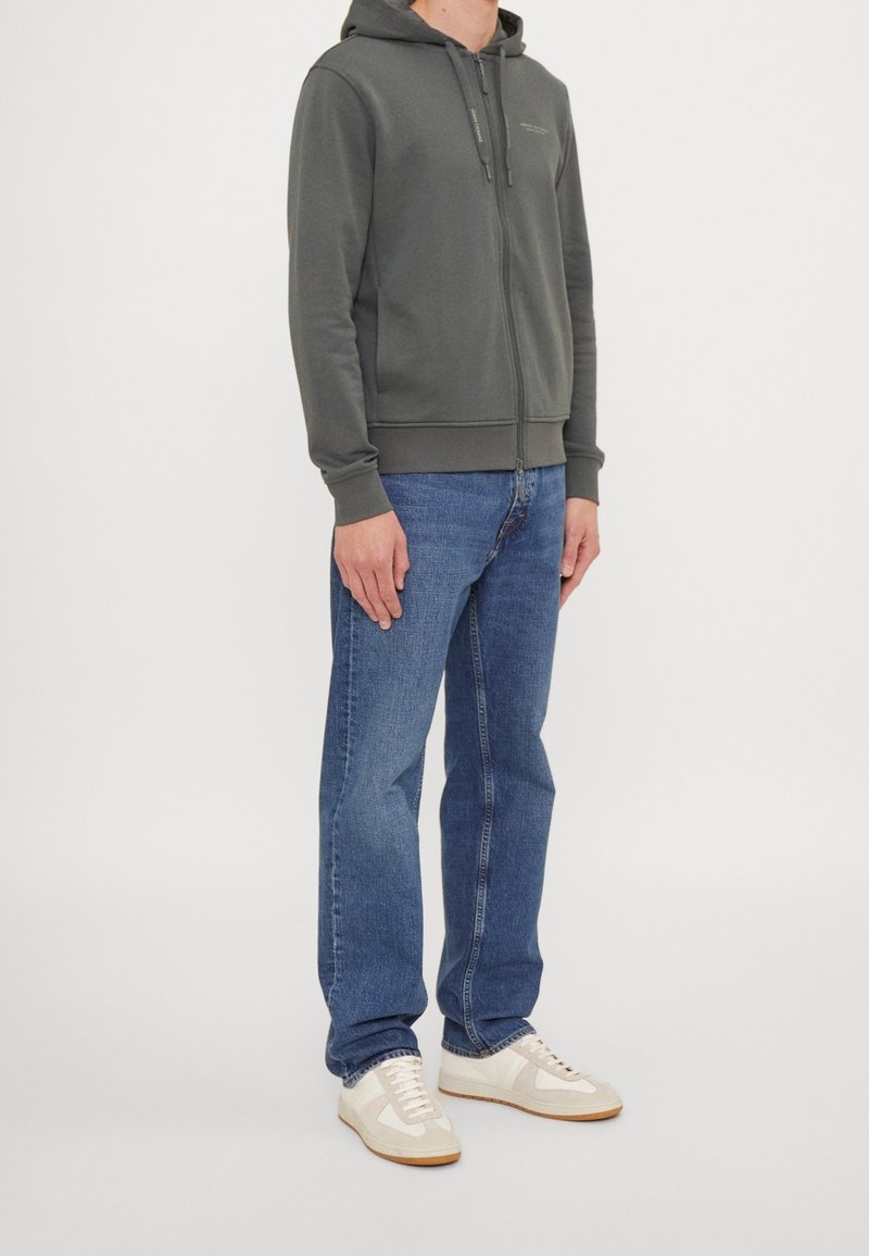 Hoodie gris zippé avec capuche, cordons, poches frontales et poignets côtelés, associé à un jean bleu et des baskets beiges. Design simple et décontracté.