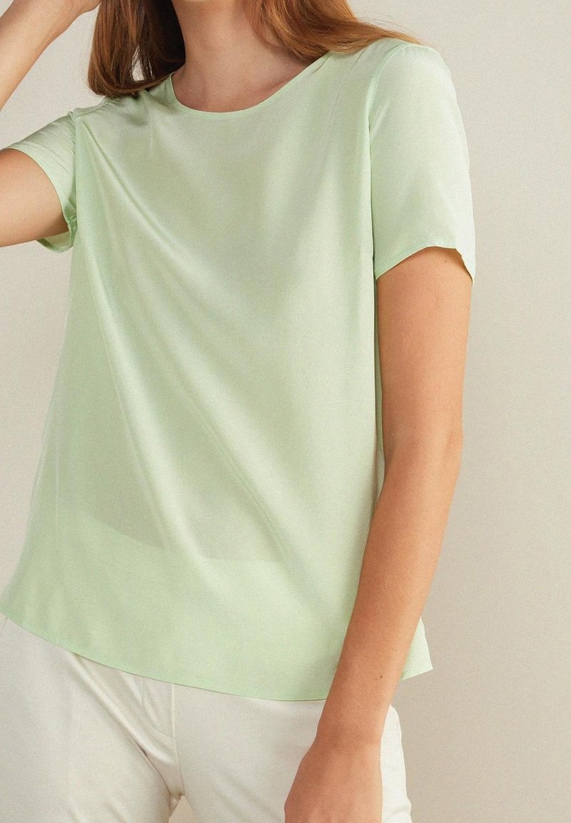 Personne portant un chemisier vert clair à manches courtes et un pantalon blanc, debout devant un fond neutre avec le bras plié près de la tête.