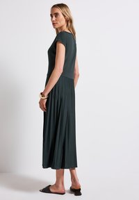 Robe vert foncé avec un haut ajusté et une jupe plissée. Manches courtes, longueur genoux, texture lisse, associée à des sandales plates noires.