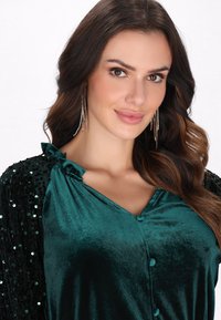 Top din catifea teal, cu decolteu în V și închidere cu buton, având mâneci decorate cu paiete negre. Materialul are o textură moale și un luciu discret.