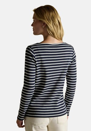 Woman with blonde hair wearing a long-sleeve navy blue and white striped top and light-colored pants, facing away from the camera.