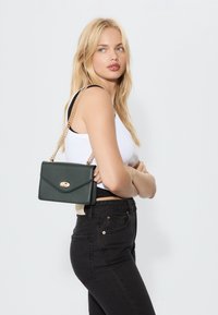 Grüne Lederhandtasche mit goldfarbenem Verschluss und Kettenriemen. Das Design ist rechteckig mit einer Umschlagklappe im Briefumschlagstil.