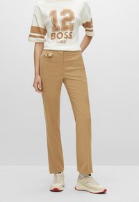 Vaaleat suorat housut, joissa on sileä pinta ja pieni etutasku, yhdistettynä valkoiseen paitaan, jossa on ruskeita grafiikoita ja raidoitusta.