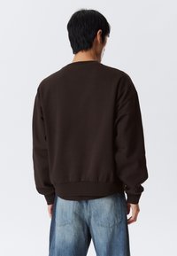 Homme tourné vers l'arrière portant un sweat-shirt à manches longues marron foncé et un jean en denim bleu clair sur un fond blanc uni.