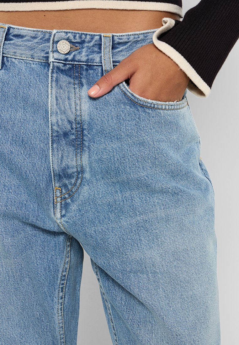 Person in hellblauen Jeans mit der rechten Hand in der Tasche, kombiniert mit einem schwarzen Langarmshirt mit cremefarbenen Manschetten.