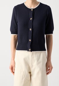 Navy knitted cardigan with short puff sleeves, ribbed collar and hem, featuring six buttons and contrasting white edges along the cuffs.