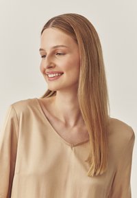Beige, leichtes Oberteil mit V-Ausschnitt und kurzen Ärmeln, das eine glatte Textur und ein einfaches Design aufweist. Die Haare sind lang und glatt.