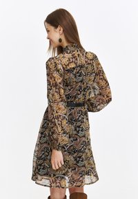 Langärmliges Kleid mit einem dunklen floralen Paisleymuster, transparentem Stoff, hohem Kragen und einem schwarzen Gürtel, der die Taille betont.