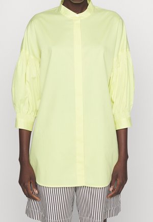 Model trägt eine hellgelbe Langarmbluse mit Puffärmeln und schwarz-weiß gestreifte Shorts vor einem einfarbigen Hintergrund.