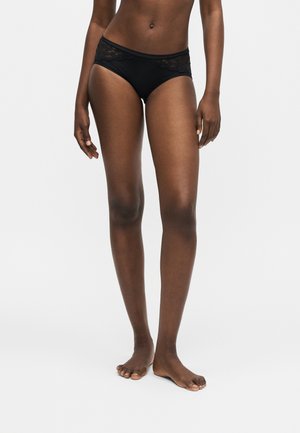 Femme portant une lingerie noire style bikini avec bordure en dentelle, debout pieds nus sur un fond clair uni.
