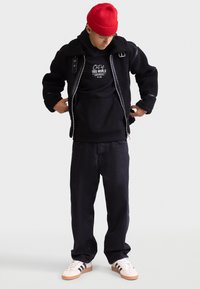 Giacca in pile nera con tessuto testurizzato, felpa nera con testo bianco, jeans scuri e sneakers bianche con strisce nere. Berretto a maglia rosso.