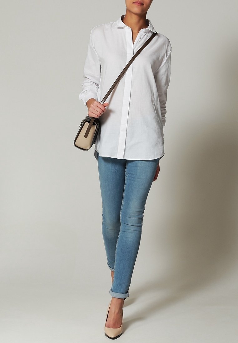 Personne portant une chemise blanche boutonnée, un jean skinny bleu clair retroussé, des talons hauts beiges et portant un sac bandoulière beige et marron.