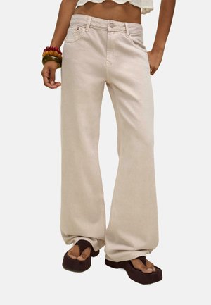 Jean large beige en tissu denim, avec une taille haute, des coutures visibles et des poches avant. Porté avec des sandales à plateforme marron.
