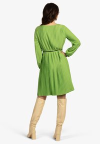 Vestido verde plisado con mangas largas y cintura definida, combinado con botas beige hasta la rodilla. Tela suave con una silueta fluida.
