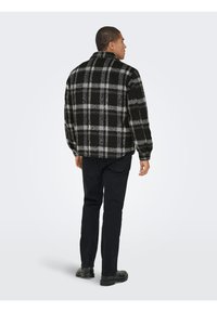 Homme debout, de dos, portant une veste à carreaux noire et grise, un pantalon noir et des chaussures noires sur un fond clair uni.