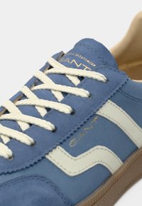 Baskets bleues GANT avec lacets blancs, bande latérale blanche et semelle en gomme, montrant des coutures détaillées et le logo de la marque sur la languette et le côté.