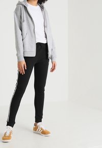 Grå zip-up hoodie med dragsko, kombinerad med svarta byxor med vita sidostraser, och beige sneakers med vita detaljer.