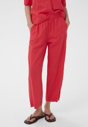 Osoba nosi voľné červené nohavice a zodpovedajúci top, stojí s rukami v vreckách, nosí tmavohnedé sandále na bielom pozadí.