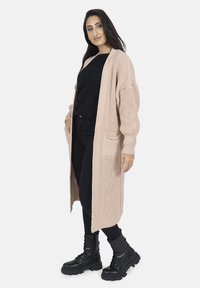 Cardigan lungo aperto in maglia rosa chiaro con maniche a costine e due tasche frontali, abbinato a un top nero e pantaloni.
