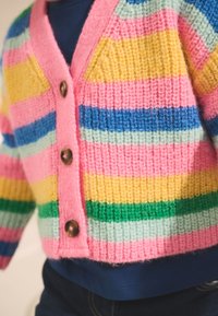 Enfant portant un cardigan tricoté coloré avec des rayures horizontales rose, jaune, bleu, vert et blanc, fermé par des boutons marron.