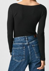 Schwarzes Langarmoberteil mit weitem Halsausschnitt, kombiniert mit hochgeschnittenen blauen Jeans, die detaillierte Gesäßtaschen und eine strukturierte Oberfläche aufweisen.