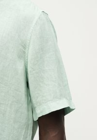 Ανοιχτό πράσινο λινό πουκάμισο με κοντά μανίκια σε άτομο με σκούρο δέρμα, εμφανιζόμενο από τον ώμο έως το μέσο του πάνω βραχίονα σε απλό φόντο.