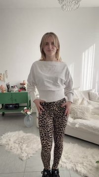 Noor naine seisab hubases elutoas, kandes valget kampsunit ja leopardimustrilisi retuuse, käed puusadel ja kergelt naeratades.