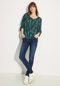 Blusa verde a fantasia con scollatura a V, abbinata a jeans skinny blu scuro e sneakers bianche con un accento verde chiaro.
