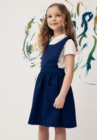 Mladé dievča s dlhou kučeravou vlasmi, ktoré má na sebe námornícku modrú zásterkovú šaty cez bielu košeľu s krátkym rukávom, stojace pri bielej stene s abstraktným umením.