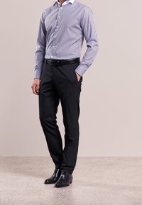 Uomo con pantaloni neri, cintura nera, scarpe eleganti nere lucidate e una camicia bianca a maniche lunghe con strisce verticali blu, in piedi all'interno.