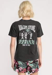 Czarny bawełniany t-shirt z grafiką tańczących szkieletów i tekstem, o luźnym kroju i krótkich rękawach. Wyświetlona plecna strona.