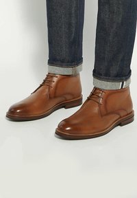 Brown leather ankle boots with a smooth finish, featuring a rounded toe, lace-up design, and a sturdy sole. Worn with dark denim jeans.