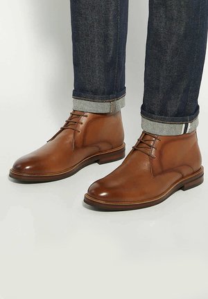 Brown leather ankle boots with a smooth finish, featuring a rounded toe, lace-up design, and a sturdy sole. Worn with dark denim jeans.